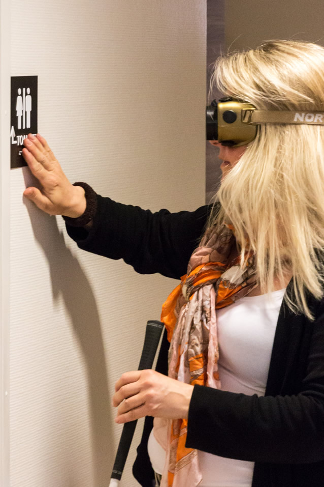 A woman in impairment glasses touching the braille and tactile letters on a sign for toilet, indoors. In the other hand, she is holding awhite cane.