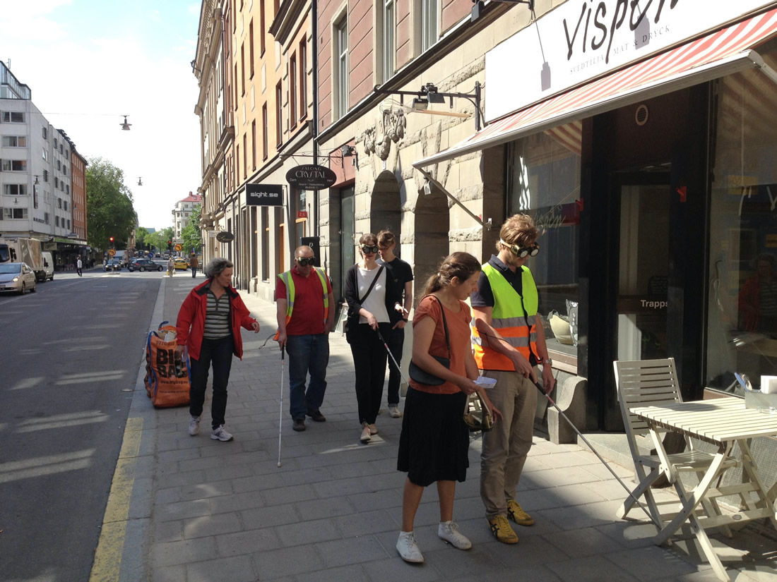 Pairs of exercise participants walking on the pavement to an outdoor café. One in each pair wearing impairment glasses and usinga white cane, the pair mate giving instructions.
