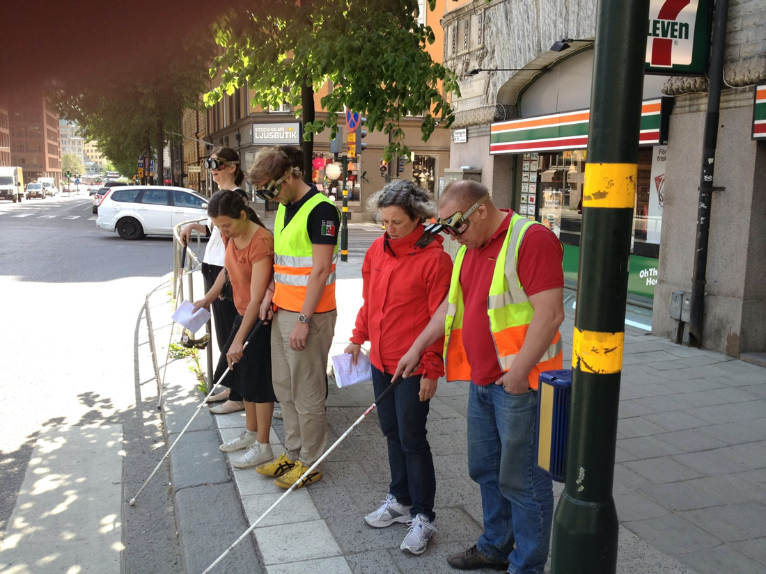 A group of exercise participants in impairment glasses and supervisors standing just in front of the street crossing, trying to feel where the street starts with the white canes.