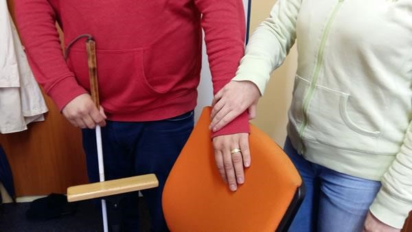 Sitting on a chair. The image shows the hand of a blind person on the back of the chair, where he has been guided with the help of his assistant.