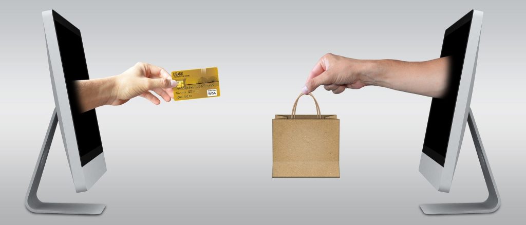 Na obrázovke sú dva počítače, z ktorých vychádzajú ruky - jedna drží platobnú kartu a druhá nákupnú tašku.