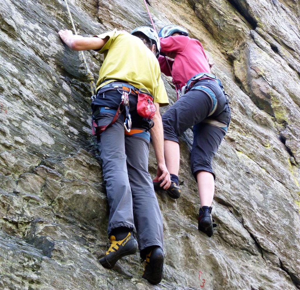 Dvaja horolezci na skale, muž pomáha žene tým, že kladie jej nohu na skalnú vyvýšeninu.