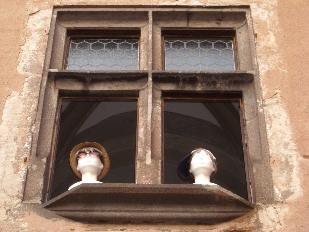 V historickom okne sú hlavy figurín. Jedna má klogúk a slnečné okuliare a druhá šiltovku. Vyzerajú akoby sa dívali z okna.