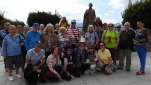 Na foto je skupinka usmievajúcich sa turistov a za nimi socha.