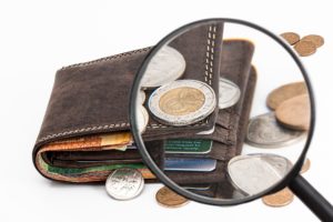 Na obrázku je peňaženka, okolo nej mince a cez je lupa.