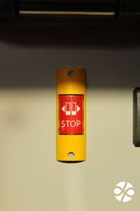 Detail tlačidla obdĺžnikového tvaru, v žltom obdĺžniku je vložený červený - uprostred je text STOP a nad ním STOP v braili.