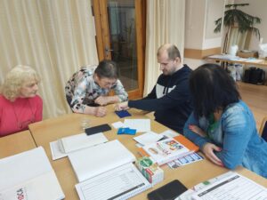Na obrázku sú okolo stola sediace účastníčky pobytu a sledujú ruky Michala Tkáčika, ako píše braila do bodkovadla.
