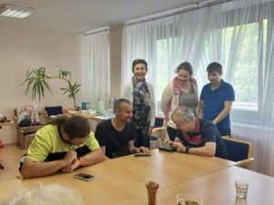 Traja hráči sedia nad mobilmi, dve ženy a mladí chlapec stoja pri nich. Mladí muž za stolom hrajú hru a všetci sa smejú.