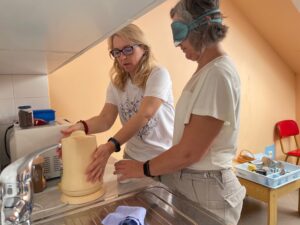 Na obrázku sú dve pracovníčky, jedna má klapky na očiach, v rukách má šálku a druhá je pri rýchlovarnej kanvici.