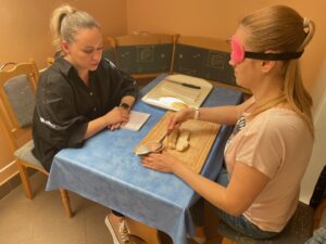 Pracovníčky sedia oproti sebe za stolom. Jedna má pred sebou poznámky a hovorí niečo na kolegyňu. Druhá sa snaží natrieť si rozpolený rožok paštétou.