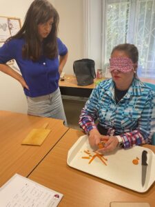 Pracovníčka sedí za stolom, na ktorom je tácka s nožíkom. Nad ňou má ruky s mrkvou, ktorú šúpe. Pri nej stojí ďalšia pracovníčka a hovorí jej, čo má robiť.