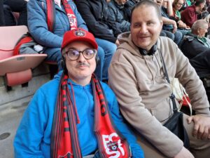 Na obrázku sedia dvaja muži v hľadiskul jeden ma na hlave červenú šiltovku FC Spartak a tiež šál na krku. Obaja sa dívajú do objektívu s jemným úsmevom. 