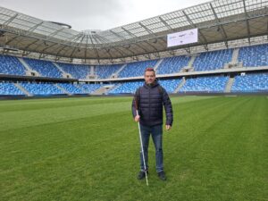 Na obrázku je uprostred hracieho poľa štadióna mladý muž s bielou palicou. Okolo neho je veľké prázdne hľadisko s modrými stoličkami.