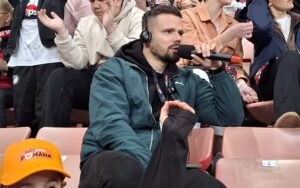 Na obrázku je vidno predovšetkým záber na moderátora, ktorý sleduje zápas, na ušiach má slúchadlá a v ruke (pri ústach) mikrofón.