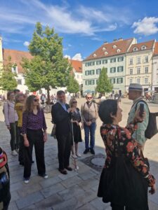 V Starom Meste je skupina turistov, niektorí majú bielu palicu, tmavé okuliare. V popredí je V. Bertrand-Vivancos, ktorá si fotí okolie. Za ňou vidno sprievodcu, ktorý skupinke niečo hovorí.
